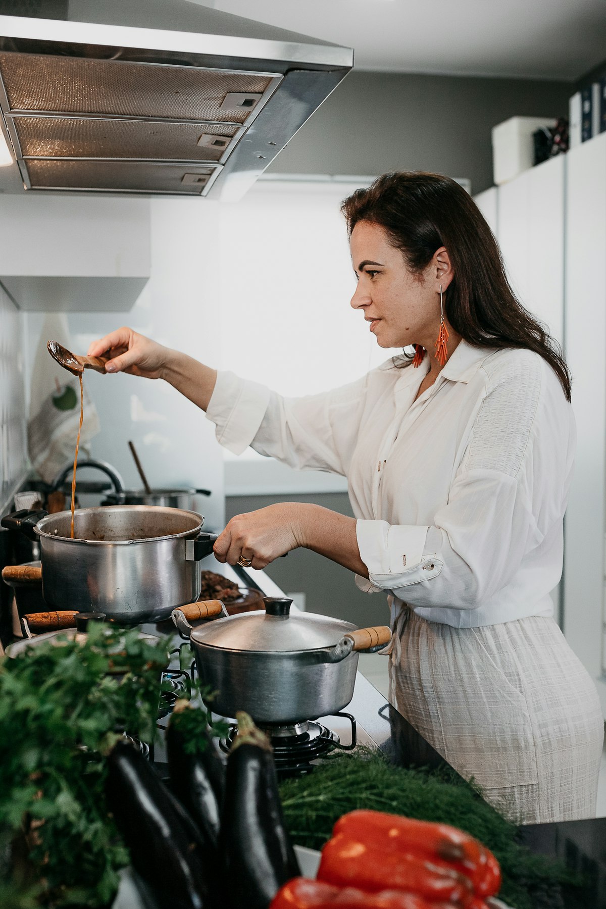 A home cook tasting a sauce from a wooden spoon at her own stove, fresh vegetables on the counter beside her
